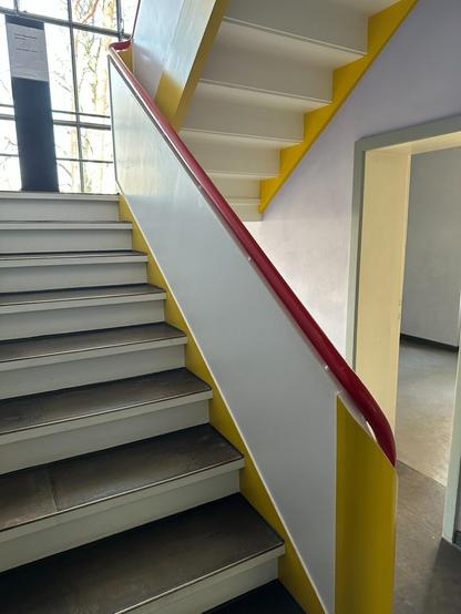 A staircase ascending. The bannisters are composed of geometric forms and painted brightly, white with red and yellow accents. A large window lets in sunlight