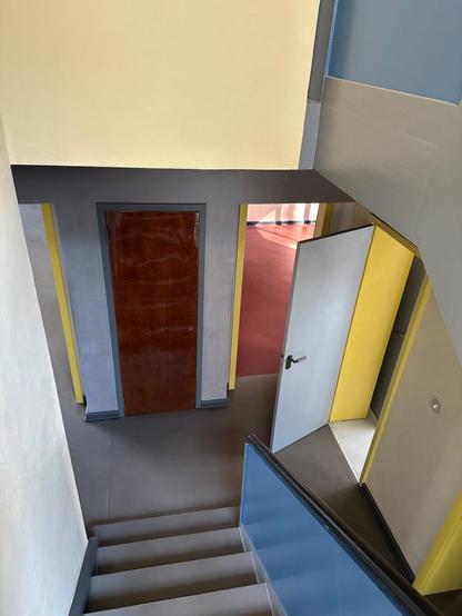 A corridor seen from a mezzanine stair landing above. Various doors are open. Surfaces are painted colourfully, with red, light yellow and various shades of blue-grey.
