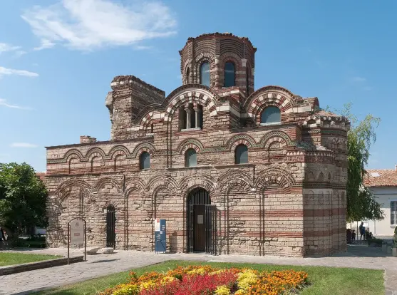 The image depicts a historic building characterized by intricate brickwork and a unique architectural style. It features a dome and several arched windows and doorways, set against a blue sky with some clouds. In the foreground, there are colorful flower beds. This type of architecture suggests it may be a church or a significant cultural site, likely from the Byzantine or early medieval period.

Image Credits: Wikimedia / Chrumps / CC BY-SA 4.0
