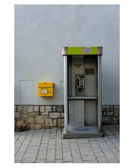Foto im Hochformat. Frontalaufnahme einer österreichischen Telefonzelle, die an einer Wand steht. Links daneben ist ein gelber Briefkasten. Die Telefonzelle ist vorne offen und hat ein grün-rosafarbenes Dach. Darin hängt ein Münzfernsprecher mit rosafarbenem Hörer. Die Wand ist einfarbig hellgrau, unten ist ein Streifen Mauerwerk. Der Fußweg davor geht leicht bergab/berrgauf, weshalb unter der Telefonzelle ein Sockel zum Ausgleich ist. Neben dem Briefkasten ist noch eine kleine Tür in der Wand, ca. doppelt so groß wie der Briefkasten.