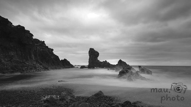 This black and white image shows a coastal scene with rocky formations extending into the water. The sky is overcast, creating a dramatic atmosphere. The water appears smooth, indicating a long exposure photograph. Rocks and pebbles are visible in the foreground.
