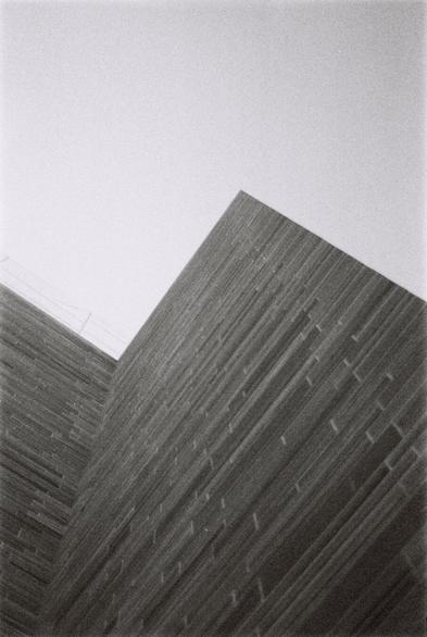 A very grainy, black-and-white photograph of a zig-zaging outer wall at the Norwegian National Museum. The wall is clad in dark slate tiles, with a top portion in larger tiles of white marble barely visible against the bright, clear sky. The wall creates a pointy up-and-down geometric shape against the sky, almost like a caricature drawing of a simple mountain range.