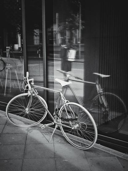 A bicycle is leaning against a reflective window of a building. The bicycle is silver-colored with white tires and a white seat. It has dropped handlebars and a kickstand. The reflection of the bicycle, a passerby in a dark coat, with a white backpack and some trees and buildings are visible on the surface it is leaning against. The ground in front of the bicycle is paved with square tiles. The image is in black and white, portrait-orientated.