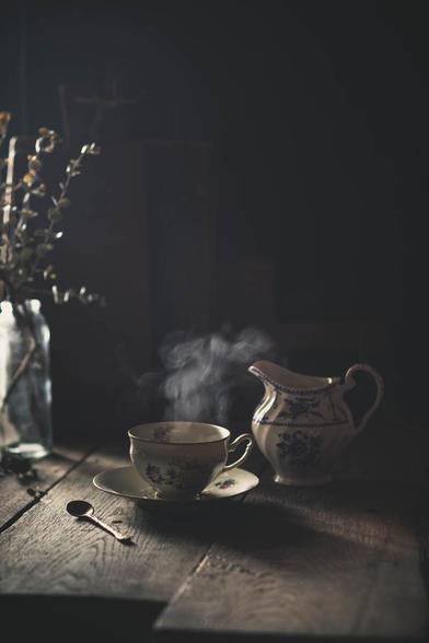 Alte Tasse, mit dampfendem Tee gefüllt. Ein Milchkännchen rechts daneben, links eine Glasvase mit trockenen Zweigen. Sonnenlicht fällt von rechts in das Bild.