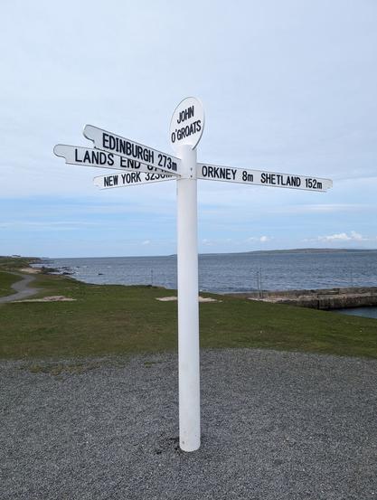 A white signpost standing with the sea in the background. The sign at the top says "John O'Groats" and the four arms show the directions and distances to: 

 (Left)
New York   3230m
Lands End   874m
Edinburgh   273m

(Right)
Orkney  8m   Shetland  152m