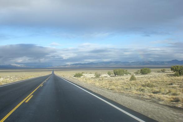 A long, straight, paved, two-lane road that disappears into the horizon where there are mountains, with mostly cloudy overhead, and sage brush off the narrow shoulders.