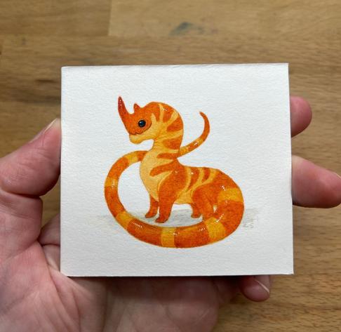 a hand holding a tiny painting of an orange dragon with darker orange stripes and a long tail and a black beady eye and two horns.