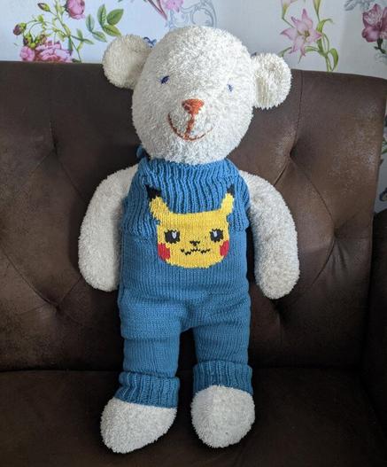 Photo of a white teddy bear standing up on the sofa wearing blue knitted overalls. There is a yellow pikachu face smiling in the middle of the chest of the overalls.