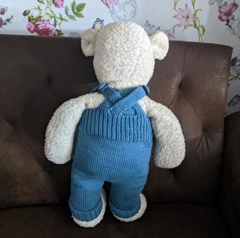 Photo of the back of the same teddy bear. You can see where the straps of the overalls are crossed over. The straps are joined with a plastic clasp and they have a long tail. The tails are too long and make it look like a bit of a mess.