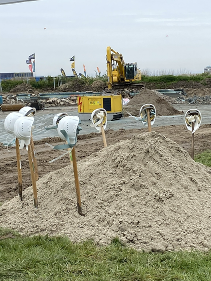 Baustelle des Freibads in Norddeichmit Bagger und viel Sand. Der erste Spatenstich wird durch 7 Spaten und weiße Bauhelme mit Schleifen symbolisiert