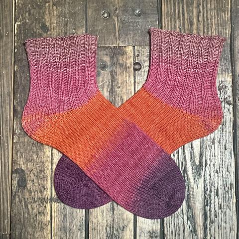 Flat-lay of a pair of hand knitted socks in shades of pink, orange, and purple against a wooden floor. The foot is all knit stitch, and the ankles are k2p2 rib knit.