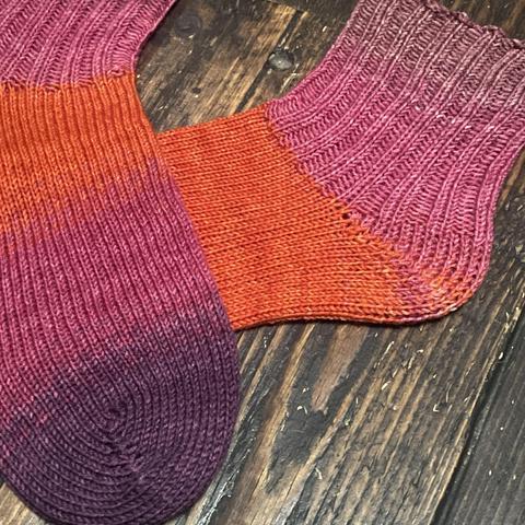 Detail of a pair of hand knitted socks in shades of pink, orange, and purple against a wooden floor. The foot is all knit stitch, and the ankles are k2p2 rib knit.