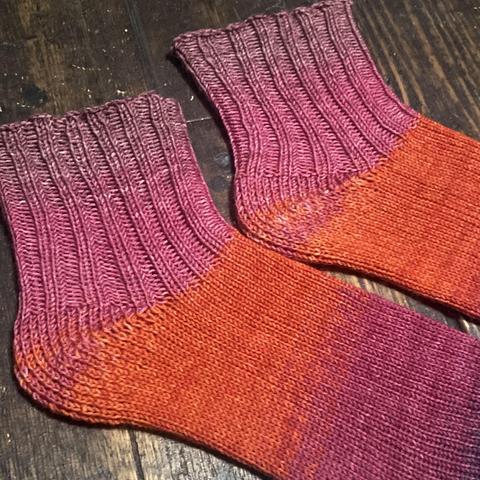 Flat-lay of a pair of hand knitted socks in shades of pink, orange, and purple against a wooden floor. The foot is all knit stitch, and the ankles are k2p2 rib knit.