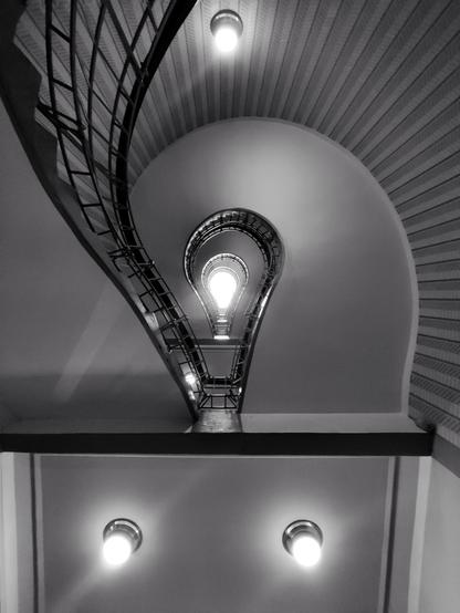 Schwarz-Weiß-Foto. Blick von unten durch ein Treppenhaus nach oben. Das Treppenhaus ist so gebaut, dass die Geländer an die Form einer Glühbirne erinnern, ganz oben fällt Tageslicht durch ein ebenfalls glühbirnenförmiges Fenster herein, so dass es aussieht, als ob die Führung leuchtet. Zusätzlich sieht man noch drei andere Lampen, zwei am unteren und eine am oberen Bildrand.