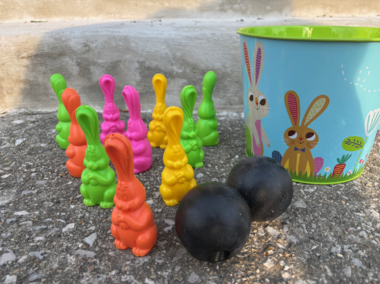 Color image of a plastic Easter bunny lawn bowling set. Image shows colorful ten plastic bunnies shaped as bowling pins, two black plastic bowling balls, and a small festive Easter metal pail for carrying the set, with bunnies painted on the pail.