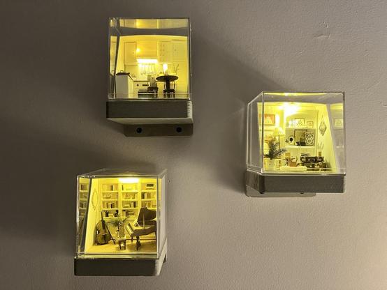 A pulled back picture of the three rooms in acrylic cubes on grey wooden shelves. Lit up, they look like impossibly tiny rooms. They are arranged somewhat haphazardly in a circle/ wonky triangle.
