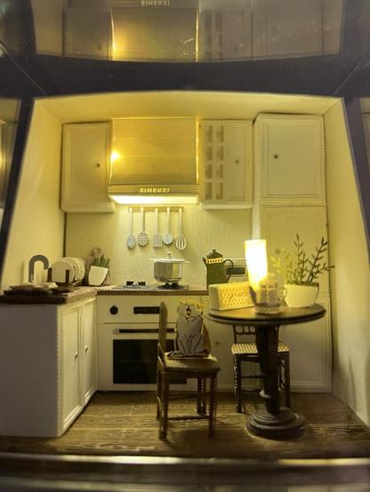 Close up of the kitchen room. There is a brown counter top, with sink, oven, range, drying rack. On the counter is a cheese board, pot on burner, and an electric tea kettle. Utensils hang behind the stove. The cupboard and range hood have details. There is a wooden table and chairs. A kitty sits on a chair yawning. On the table is a teapot, light, plant, and box. There is wood grain on the floor. The range hood is also lit.