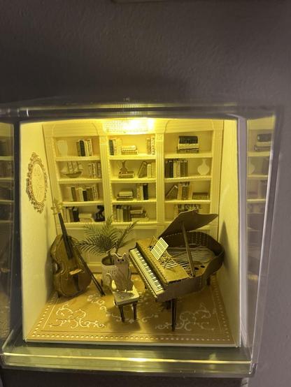 Overhead view of the music room. The back wall is a bookshelf, tastefully arranged with books, vases, and a ship. The wall has a gilded, decoratively framed rose oval piece of art. There is a baby grand piano with a piece of sheet music. A cat sits on the bench. A chelo or base sits upright with a bow leaning against it. A potted fern is also on the parquet floor. It is lit by an overhead light. Most items are white, off white, tan, or brown. There are gold and green accents.