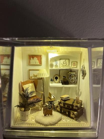 A tiny living room scene. The walls are white and have art and a mirror hung on them. A white circle light fixture illuminates it with soft light. It has a full curio, a leather chair, a ficus plant, suitcases stacked with books and quills on them and a kitty walking across a fluffy carpet.