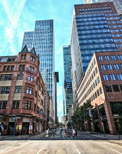 Foto einer Kreuzung in Frankfurt. Ein paar Menschen passieren die Straße, Hochhäuser mit Glasfassaden säumen selbige. Vorne ein etwas älter anmutendes Gebäude, was zumindest ein wenig Kontrast zu den moderneren Bauten bietet.