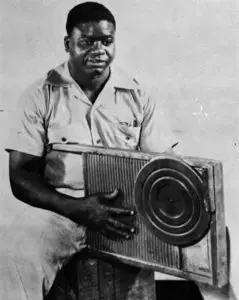 Photographie en N&B du bluesman noir américain Washboard Sam en combinaison claire avec son washboard entre les jambes