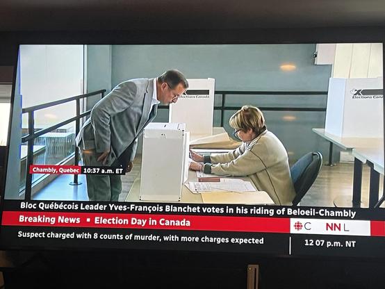 The leader of the Bloc party is shown in a gray suit, leaning over table where a woman is preparing his ballot. There is an elections candidate voting booth behind him. The ballot box is beside him on the table.