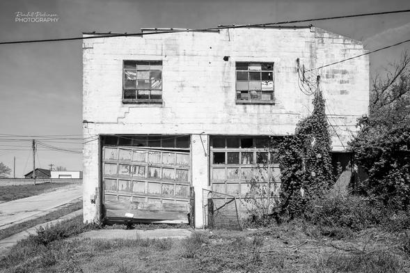 An old, abadoned concrete block building.  The lower level has two garage entrances with doors that are out of alignment and have some broken windows.  The upstairs has two, 12-pane windows.