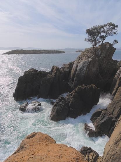 Coastal cliffs at right drop to a breaking wave, turbulent whitewater and spray surrounding an outcrop below.  Small trees cling to the rocks behind.  Islands in the bay, hazy November sky.