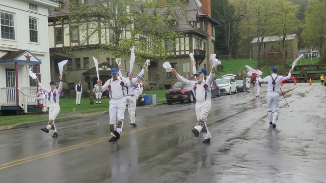 Six morris dancers with white shirts and trousers and grey suspenders, decorated with red and blue ribbons, are  in a street that is wet from recent rain. Behind them are more morris dancers, watching. At the back is a large Tudor revival inn.
