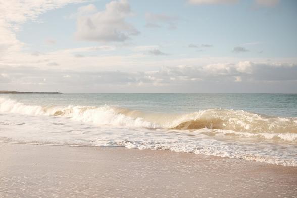 Wide shot composition of a beach scene. A singular wave dominates the midground, curling and breaking near the shore, creating bubbly, foamy white water. The foreground is a light tan sandy beach with small ripples of water. The ocean, depicted with a horizontal teal and light brown gradient, meets the sky on the horizon with a breakwater and small lighthouse visible. The sky, a soft blue with scattered, fluffy white clouds. Soft, natural light is present, with high-key lighting.