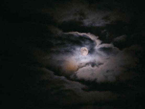 Foto vom Mond, hinter einer leicht geöffneten Wolkendecke, die vom Mondlicht angestrahlt wird.
