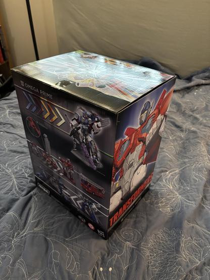 Illustrated box for Haslab Transformers Omega Prime. The back of the box illustrates the included figures in multiple modes: Omega Prime with his sword and stand; Optimus Prime as a robot, Super robot, and fire truck; and Ultra Magnus in car carrier and robot modes. The side of the box is an illustration of Optimus Prime lifting his fist.