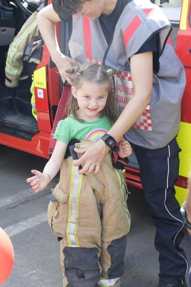 A little girl with two pigtail braids in her hair in a green t-shirt with a rainbow, up to her armpits in a seriously oversized pair of bunker pants. A female firefighter in navy blue station wear and a grey vest with red lettering is adjusting her suspenders. In the background a red van with hi viz battenburg pattern.