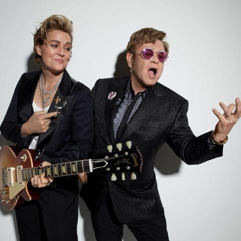 On the left, a white woman with an auburn fauxhawk and a red sunburst guitar smirks at the person standing next to her: an older man with tinted sunglasses, gesturing as he sings