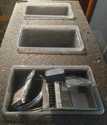Three plastic reservoirs in a block of styrofoam, one reservoir has some parts in it such as a power supply, some electronic parts in boxes and a plastic transparent tube.