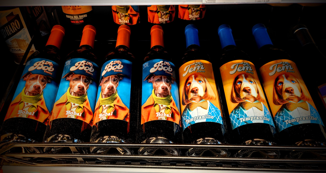 In einen Supermarkt in einem Weinregal liegen Rotweinflaschen. Auf den Etiketten sind Hunde in Kleidung abgebildet (Fotomontagen). Entsprechend den Reebsorten heißen die Mr Bob und Mr. Tem
