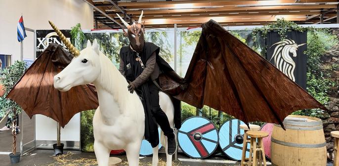 A person in a dragon costume with brown wings is riding a white unicorn statue. The background features decorative elements including green foliage and shield designs.