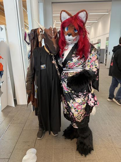 Two individuals in costumes pose together. One is wearing a mask resembling a dragon, paired with a dark outfit. The other is in a fluffy fox costume with large ears and red hair, dressed in a floral kimono. The background features a