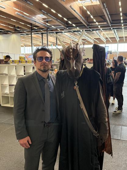 A man in a suit and sunglasses stands next to a person in a dragon-like costume with a mask and cloak. The background shows a convention setting with attendees and displays.