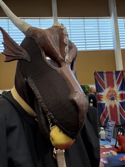A person wearing a detailed, animal-themed mask resembling a dragon, featuring horns and textured features, is holding an apple in their mouth. The background includes a visible British flag and various items on display, suggesting a convention or themed event setting.