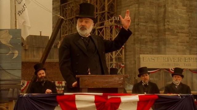 A man in an 1880s top hat gesturing at a large structure behind him. He’s standing next to a sign that says “East River Bridge”