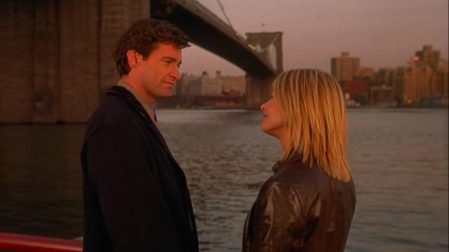 A handsome man and woman in contemporary clothes stand at the Brooklyn Bridge, gazing at each other