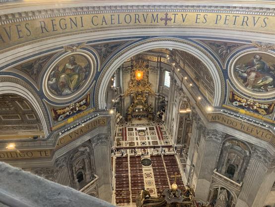 Blick von der Galerie unter der Kuppel des Petersdoms in die Tiefen des Kircheninneren. Das Foto ist durch eine Plexiglasscheibe aufgenommen, dahinter verläuft ein Handlauf. Darauf hat sich eine dicke Schicht Staub angesammelt, welche mangels Zugang nicht so einfach gewischt werden kann.