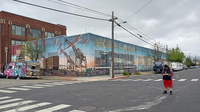 A building with a wraparound mural on two sides, showing a train and the port.  There's a colorfully decorated RV on the street.
My kid is taking a picture with a new (to him) Yashica TLR.