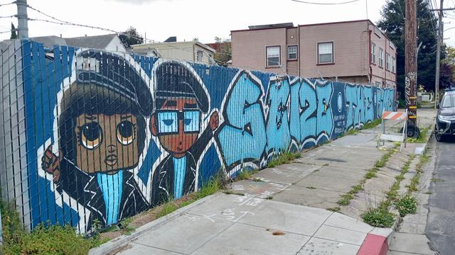 Mural on a chain-link fence with wooden slats.  It shows two militant Oaklanders in a cartoonish style.