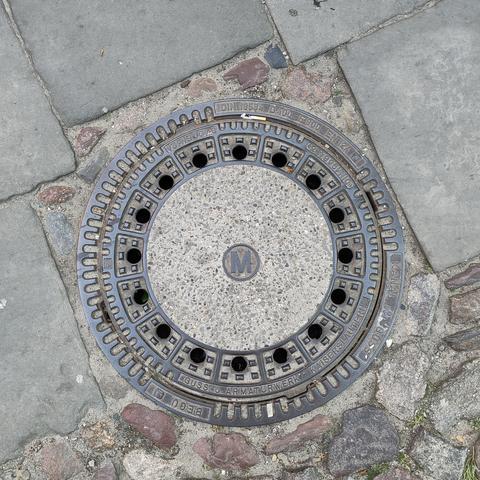 Ein runder Schachtdeckel, in dessen Zentrum alleinstehend der Buchstabe M steht.