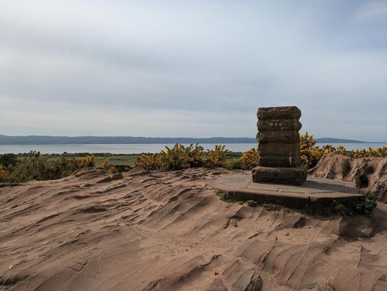 A stone monument stands on a rocky terrain overlooking a distant body of water and hills.