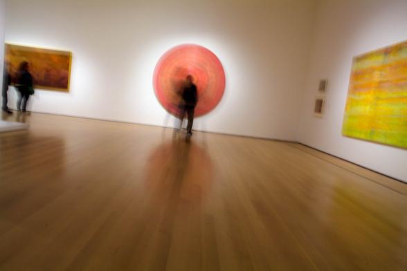 A wide, time-exposure view of one of the galleries at the Museum of Modern Art's Jack Whitten retrospective. Three large-scale pieces are visible as are two smaller ones in a corner. The composition forces the eye to the center of the frame where a blurry figure vibrates side-to-side, centered before a circular composition in the artist's unique abstract-expressionist style.