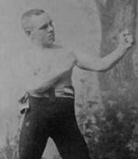 Ein Boxer in Sportkleidung steht in der Ausgangsposition für einen Schlag, mit einem leicht angespannten Gesichtsausdruck. Im Hintergrund sind unscharfe, vegetative Elemente zu sehen. Das Bild ist in Schwarzweiß gehalten.