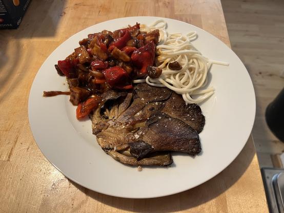 Ein Foto von dem flach gepressten und goldbraun angebratenen Austernpilzsteak, einem Haufen glänzende Paprika, Tempeh, Zwiebelstreifen in rot gläzender Sauce und einer ähnlichen Menge hell weißer Udons.